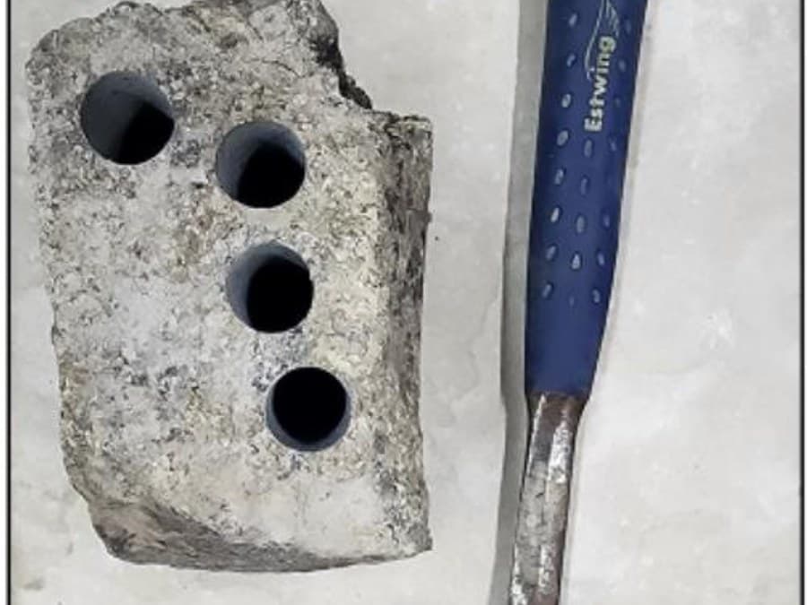 Rock block sample with drilled test holes beside a geological hammer for scale
