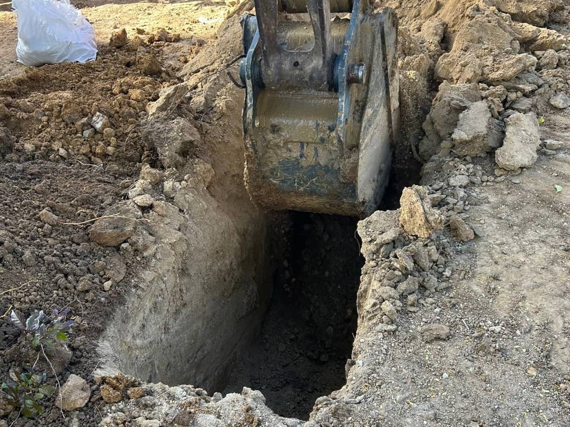 Excavator bucket above a freshly dug test pit exposing the soil profile